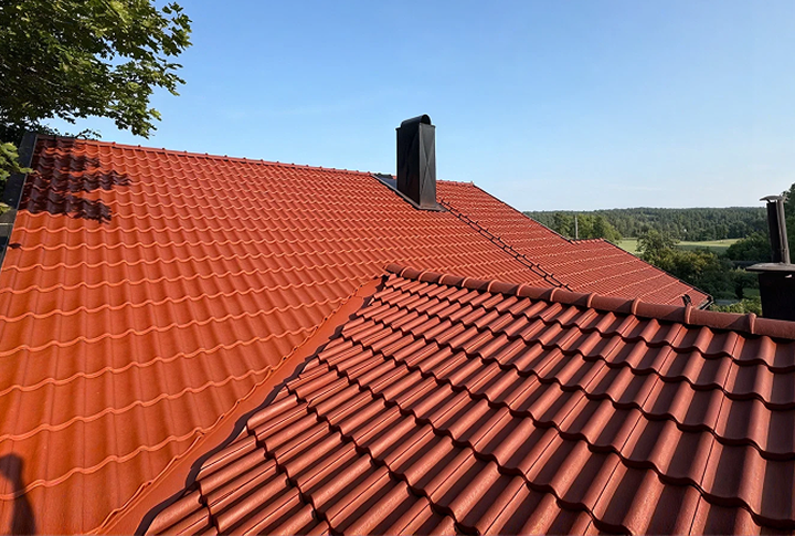 Et skråtak dekket av rød-oransje, profilerte takstein – muligens sammenføybare tegl- eller betongstein – med mørke piper mot en klar blå himmel.