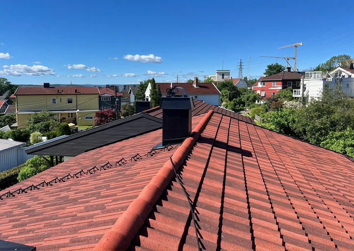 Tak dekket av røde takstein i betong eller tegl med overlapp, med en mørk pipe og en kabel som går på tvers av takflaten.