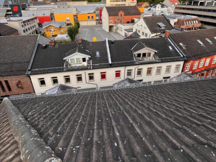 Sett ovenfra, med blikket rettet ned over et mørkt tak av bølgeformet skifer eller fibersement-takstein, mot et bybilde med bygninger i tradisjonell europeisk stil.