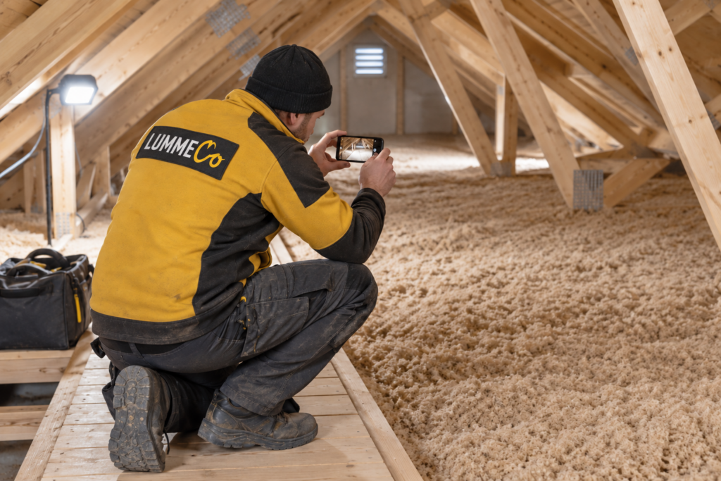 Person på loftet som inspiserer og fotograferer løsisolasjon av cellulose mellom gulvbjelkene.