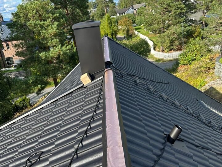 Skrått boligtak med mørke metallplater i taksteinprofil, svart skorstein og sikkerhetsline.