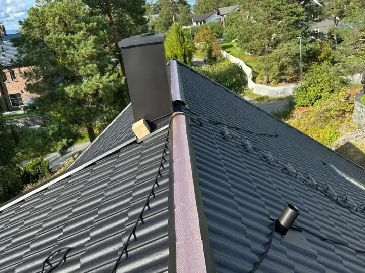 Skrått boligtak med mørke metallplater i taksteinprofil, svart skorstein og sikkerhetsline.