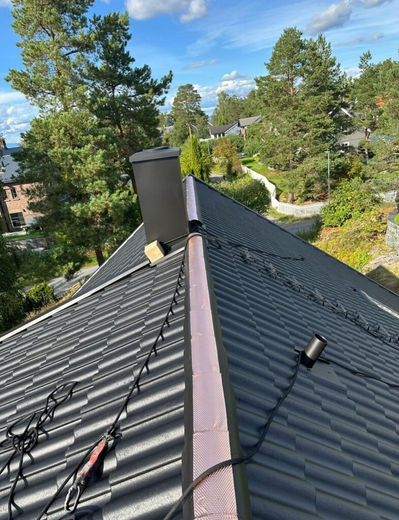 Skrått boligtak med mørke metallplater i taksteinprofil, svart skorstein og sikkerhetsline.