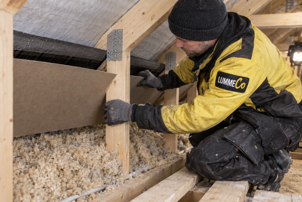 Arbeider som legger løsisolasjon mellom trebjelkene i takkonstruksjonen på et loft.