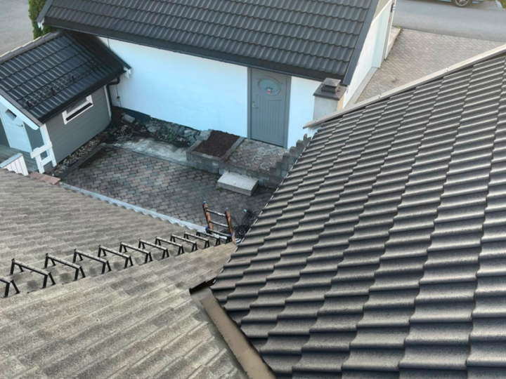 Mørkt tak med en stige plassert langs takflaten, med utsikt mot et asfaltert gårdsområde ved en hvit og grå bygning.